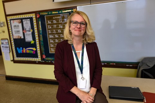 A blonde woman wearing glasses, a white shirt and suit with a lanyard around her neck.