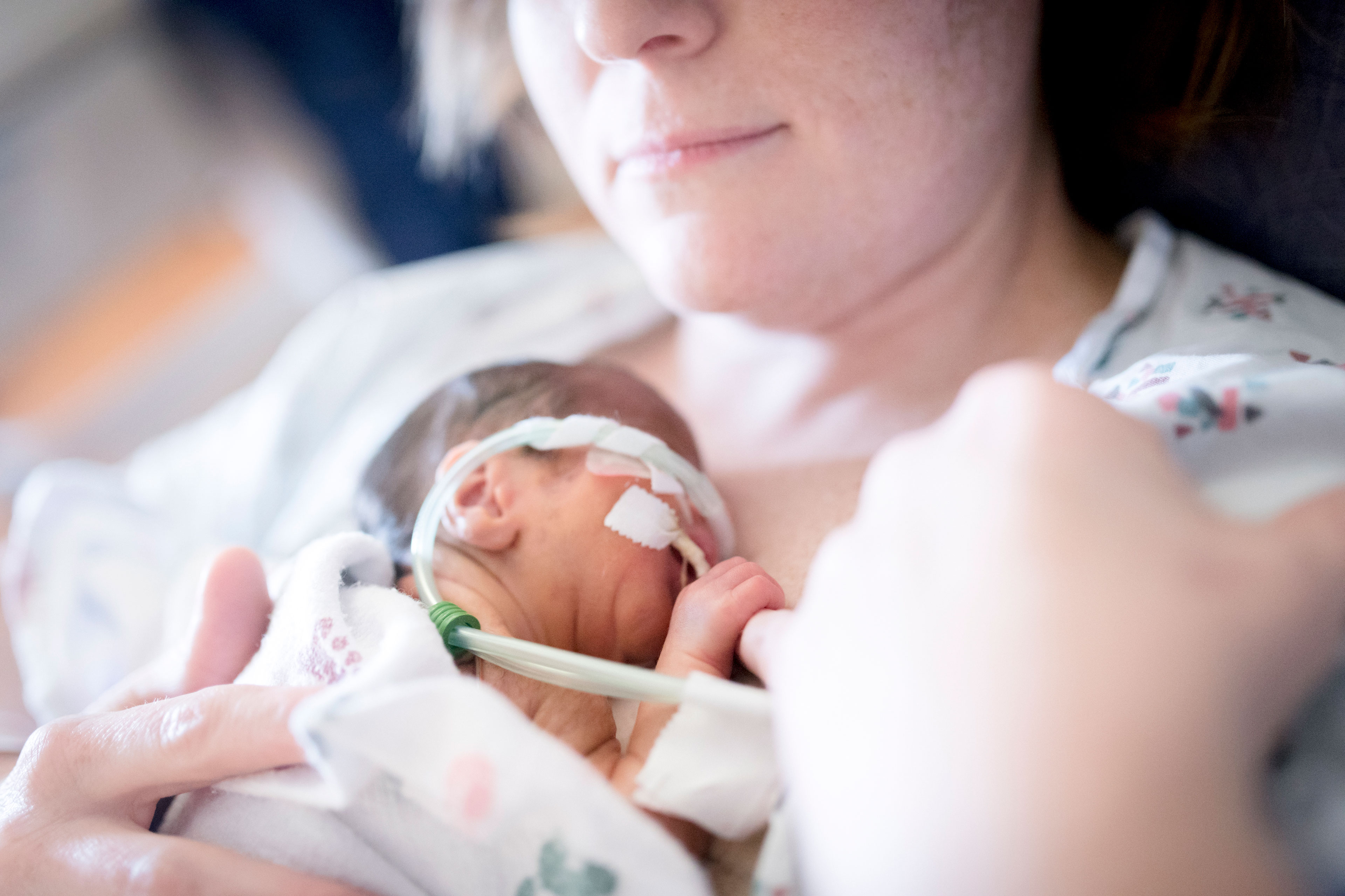 A woman holds a small newborn baby to her chest. The baby holds the woman's pinky finger.