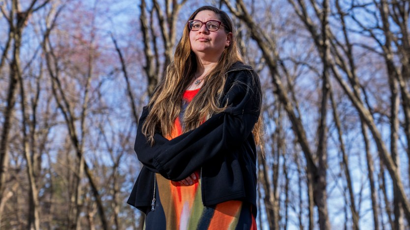 A woman stands outside in a wooded area.