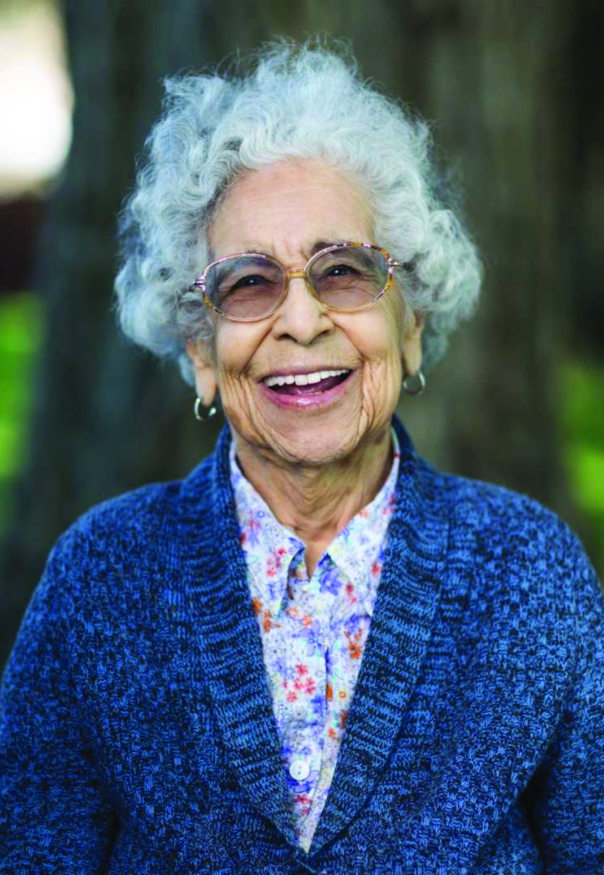 A woman with white hair and glasses, wearing a blue sweater and a floral shirt, smiles for a portrait